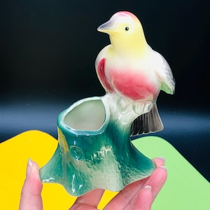 Puede incluir: Una maceta de pájaro de cerámica vintage. El pájaro tiene colores amarillo, rojo y gris. La maceta es un tocón de árbol verde con una pequeña abertura para plantas. El pájaro está posado en el tocón.