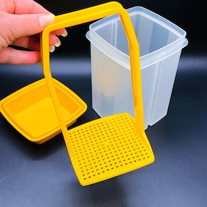 Puede incluir: Un juego de recipientes de almacenamiento de alimentos de plástico amarillo. El juego incluye un recipiente cuadrado, un inserto amarillo ranurado y una tapa. El recipiente es transparente, y el inserto y la tapa son de color amarillo brillante. Diseñado para almacenar alimentos.