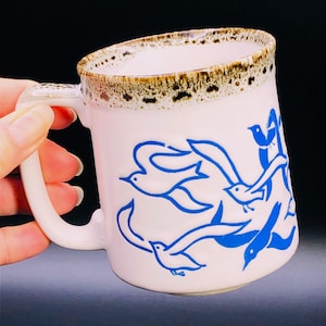 May include: White ceramic mug with a curved handle, featuring a blue bird design. The rim has a brown speckled pattern. The mug is held by a hand against a dark background. A decorative piece for the kitchen.