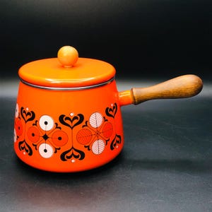 Peut inclure: Une casserole en émail orange avec une poignée et un couvercle en bois. La casserole présente un motif floral noir et blanc décoratif autour de la base. Le couvercle a un petit bouton en bois.