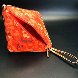 May include: An open, orange cosmetic bag with a floral pattern in shades of orange, gold, and brown. The bag has a gold zipper and a gold wrist strap. The interior is red.