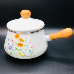 May include: White enamel saucepan with a wooden handle and lid. The saucepan has a floral design with yellow, orange, pink, blue, and green flowers. The brand name "Saltina" is printed on the bottom of the saucepan.