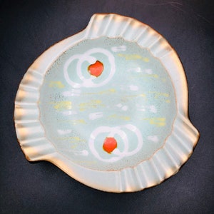 May include: A ceramic ashtray with a pale blue interior and a ruffled edge. The interior features white and yellow abstract designs, with two orange circles. The rim has a gold-coloured trim.