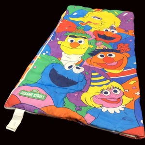 May include: A vibrant Sesame Street sleeping bag with characters including Cookie Monster, Big Bird, and Elmo. It has a white zip and the Sesame Street logo. The background is a mix of bright colours.