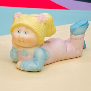 May include: A vintage Cabbage Patch Kids figurine in a lying down pose. The doll has blonde hair with pink bows, blue eyes, and is wearing a pink outfit with blue sleeves and blue shoes. The figurine is made of ceramic.