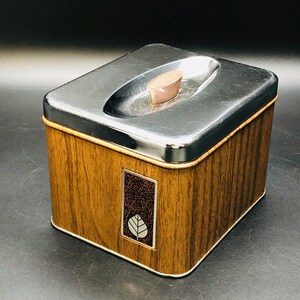 Puede incluir: Una caja cuadrada de madera con una tapa plateada y un diseño de hoja. La tapa tiene una pequeña manija ovalada.