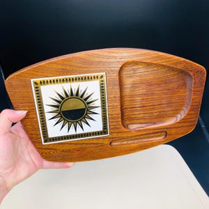 Op de afbeelding: Een vintage houten serveerschaal met een decoratief tegelinzetstuk met een zonne-uitbarstingontwerp in goud en zwart. De schaal heeft een ingebouwd compartiment en een groef, met een warme houtnerf afwerking.