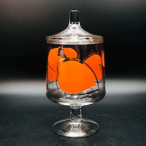 May include: A clear glass candy dish with a lid. The dish has a stem and a round base. It's decorated with orange leaf shapes outlined in black and gold. The lid has a black finial.
