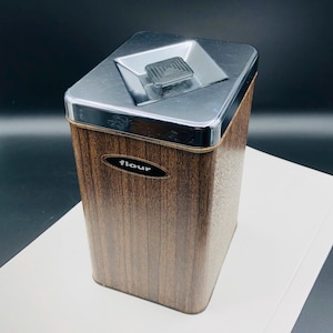 May include: A brown and silver vintage flour canister with a woodgrain pattern and a silver lid with a push-button top. The canister has the word "flour" printed on the front.