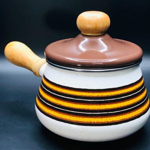 Peut inclure: Une casserole vintage en émail blanc avec un couvercle et une poignée marron. La casserole présente des rayures horizontales dans les tons orange, jaune et marron. La poignée et le bouton du couvercle sont en bois.