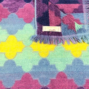 May include: A colorful, patterned towel with a geometric design in shades of purple, pink, blue, yellow, and green. The towel has a fringed edge and a label that reads "Camtex".