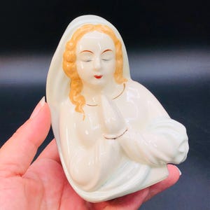May include: A cream-colored ceramic figurine depicting a woman with closed eyes and clasped hands. She wears a white veil and robe, with blonde hair and red lips. The figurine is a religious item.