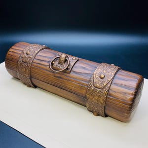 May include: A cylindrical wooden box with a rustic design. The box has a dark brown finish, with bronze-coloured metal bands and a ring handle. The box has a textured, wood-grain appearance.