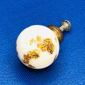 Op de afbeelding: Witte keramische kastknop met goudblad accenten. De knop heeft een ronde vorm en is bevestigd aan een metalen schroef. De hardware staat tegen een blauwe achtergrond.