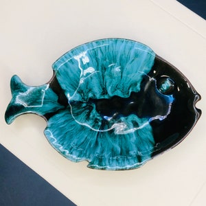 May include: A ceramic fish-shaped serving dish with a glossy finish. The dish features a striking color palette of teal and black, with a marbled effect. The fish has a detailed eye and fin, adding to its realistic appearance. This decorative dish is ideal for serving appetizers or snacks.