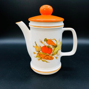 Op de afbeelding: Een witte keramische koffiekan met een oranje deksel en handvat. De kan heeft een kleurrijk ontwerp van groenten, waaronder maïs, wortels en tomaten.