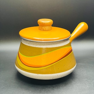 May include: A vintage orange and yellow enamelware saucepan with a wooden handle and lid. The pot features a decorative wave pattern in shades of orange, yellow, and brown. The lid has a wooden knob. The pot is approximately 6 inches in diameter.