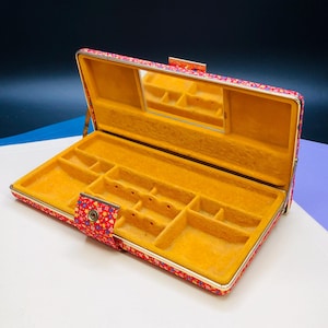May include: Open red floral patterned jewelry box with a gold-coloured trim. The interior is lined with yellow fabric and features a mirror and compartments for storing jewellery. The box is rectangular and has a clasp closure.