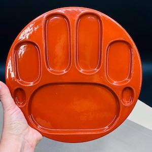 May include: A round, orange serving tray with a paw-shaped design. The tray has a glossy finish and features five oval compartments and one large, oval compartment. The tray is held by a hand.