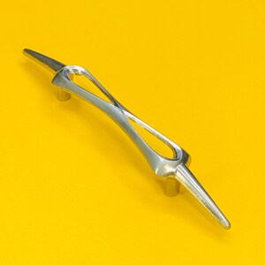 May include: A silver cabinet pull with an unusual, open design. The handle has a central, elongated oval shape with pointed ends. The pull is mounted on two small, cylindrical posts. The background is a bright yellow.