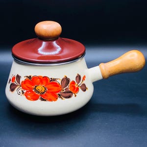 Puede incluir: Cacerola de esmalte crema con un diseño floral en naranja y marrón. La olla tiene una tapa roja oscura con un pomo de madera y un asa de madera. Un artículo de cocina vintage.
