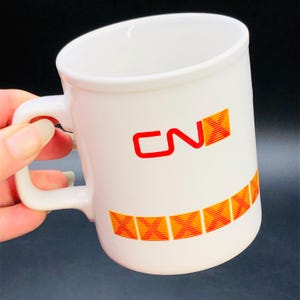 May include: White ceramic mug with a curved handle. The mug features the red letters "CN" and a series of orange and yellow geometric designs. The mug is designed for hot beverages.