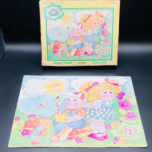 May include: A 60-piece jigsaw puzzle featuring two Cabbage Patch Kids dolls. The dolls are dressed in colorful outfits and are surrounded by flowers and mushrooms. The puzzle box is also shown in the image. The box is blue and white and features the Cabbage Patch Kids logo.