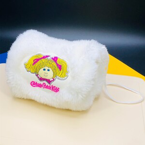 May include: A white, plush Cabbage Patch Kids purse with a cartoon graphic of a blonde girl with pink bows. The purse has a white strap and is set against a colorful background.