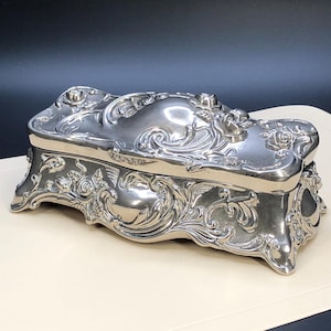 Puede incluir: Una caja rectangular de metal plateado con un diseño en relieve de volutas y motivos florales. La tapa y los laterales presentan patrones intrincados, y la caja se apoya sobre cuatro patas decorativas.