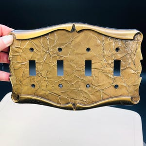 May include: A decorative light switch plate with four switch openings. The plate has a textured, gold-coloured surface with a bronze-coloured trim. The plate is held by a hand.