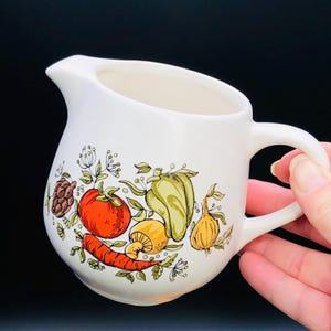 May include: White ceramic creamer with a colourful vegetable design. The design includes a red tomato, green pepper, yellow onion, orange carrot, and brown mushroom.