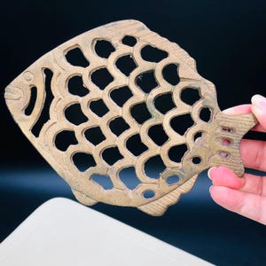 May include: A brass-colored, fish-shaped trivet with a cutout design. The trivet features a detailed scale pattern and a fin-shaped handle. The fish's eye and mouth are also visible. The trivet is held by a hand against a dark background.
