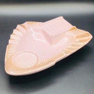 May include: A vintage pink ashtray with a unique design. The ashtray features a rectangular rest, a circular indentation, and a textured rim with a brown trim. The ashtray is made of ceramic and has a glossy finish.