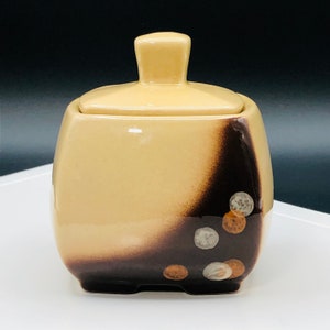 May include: A ceramic sugar bowl with a lid. The bowl is a light tan color with a dark brown gradient on one side. The design includes small, circular accents in silver and copper tones. The lid has a small, square handle.