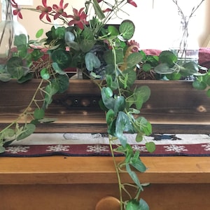 May include: A wooden planter box with a dark brown stain. The planter is filled with green leafy plants and red flowers. The planter is sitting on a wooden table with a red and white patterned runner.