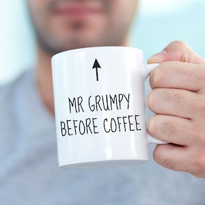 Puede incluir: Taza de café de cerámica blanca con el texto "MR GRUMPY BEFORE COFFEE" en negro, y una flecha negra que apunta hacia arriba. La taza está sujeta por una persona.