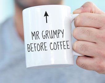 Może przedstawiać: Biały ceramiczny kubek z czarną strzałką skierowaną w górę nad napisem "MR GRUMPY BEFORE COFFEE" wydrukowanym prostą czcionką bezszeryfową. Kubek posiada standardowe ucho oraz gładki, cylindryczny korpus o regularnym kształcie.