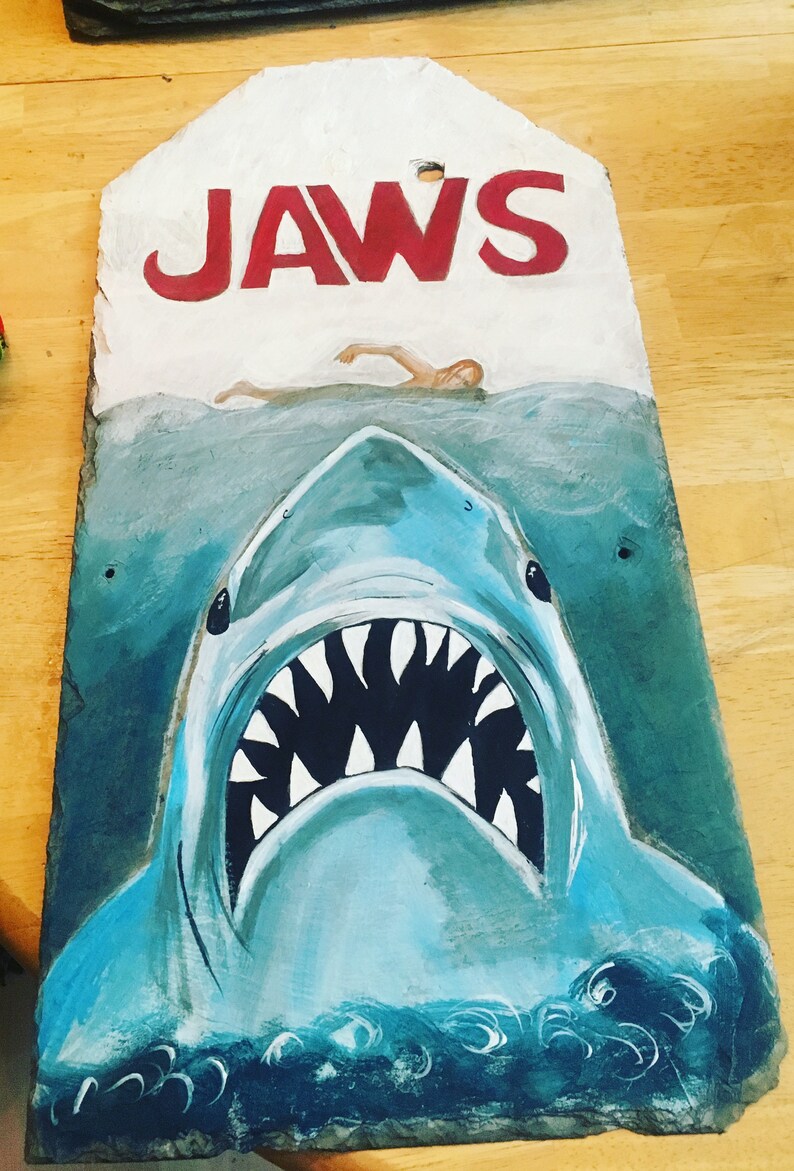 May include: A hand-painted slate sign with the word "JAWS" in red letters. The sign depicts a large shark with its mouth open, revealing sharp teeth, and a person swimming in the water above the shark.