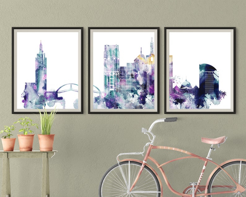 May include: Three framed watercolor paintings of cityscapes in shades of purple, blue, and green. The paintings are hung on a light gray wall.