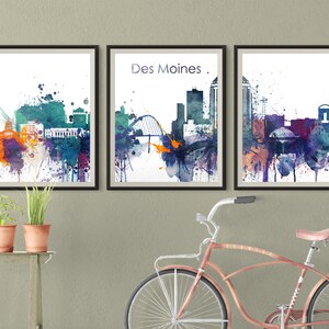 Könnte beinhalten: Drei Aquarelle der Skyline von Des Moines. Die Bilder sind in schwarzen Rahmen und hängen an einer hellgrauen Wand. Das mittlere Bild trägt den Text "Des Moines." in Schwarz.