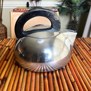 May include: A stainless steel teapot with a black handle. The teapot has a rounded body and a spout for pouring. It sits on a bamboo mat. The teapot is a kitchenware item.
