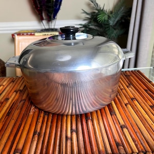May include: A shiny, silver-colored cooking pot with a black handle on the lid. The pot has two side handles and is sitting on a brown bamboo mat. The pot is round and has a lid.