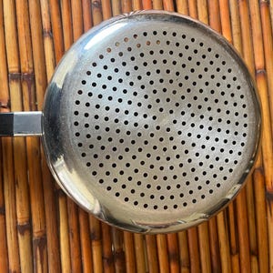 May include: A stainless steel strainer with a perforated bowl and a long handle. The strainer is round and has numerous small holes for draining liquids. The handle is black and attached to the bowl with a silver metal piece. The strainer is designed for kitchen use.