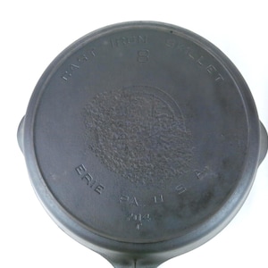 May include: A dark gray cast iron skillet, viewed from the bottom, with the words "Erie PA USA" and "8" visible. The skillet has a handle and a circular design in the center.