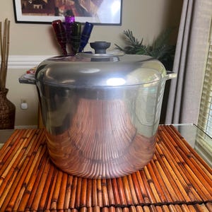 May include: A large, shiny, silver-colored cooking pot with a lid and a black handle. The pot is sitting on a bamboo mat. The pot has two side handles and a round lid with a black knob.