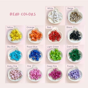 Peut inclure: Collection de perles alphabet de diff&eacute;rentes couleurs : blanc, beige, jaune, orange, rouge, noir, bleu ciel, bleu roi, vert clair, vert fonc&eacute;, rose b&eacute;b&eacute;, rose, rose fonc&eacute; et violet. Les perles sont pr&eacute;sent&eacute;es dans de petites coupelles en forme de coquillage.
