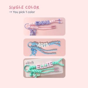 Peut inclure: Trois porte-cl&eacute;s personnalis&eacute;s avec les noms "EMMA", "ARIA" et "ELLIE". Chaque porte-cl&eacute;s pr&eacute;sente une combinaison de couleurs diff&eacute;rente, notamment rose, bleu et vert. Le texte "SINGLE COLOR" et "You pick 1 color" est en haut.