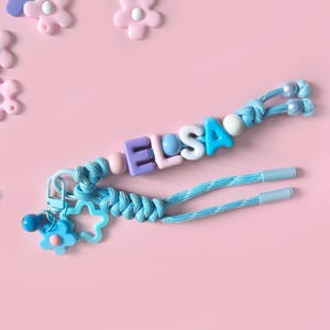 Peut inclure: Un porte-cl&eacute;s bleu clair avec le nom "ELSA" &eacute;pel&eacute; avec des perles de lettres color&eacute;es. Le porte-cl&eacute;s comporte un charm en forme de fleur et d'&eacute;toile, sur un fond rose avec des ornements en forme de fleur. Mesure environ 15 cm.