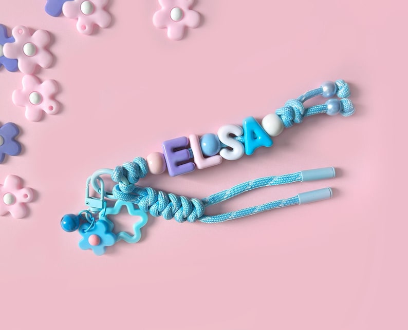 Peut inclure: Un porte-cl&eacute;s bleu clair avec le nom "ELSA" &eacute;pel&eacute; avec des perles de lettres color&eacute;es. Le porte-cl&eacute;s comporte un charm en forme de fleur et d'&eacute;toile, sur un fond rose avec des ornements en forme de fleur. Mesure environ 15 cm.