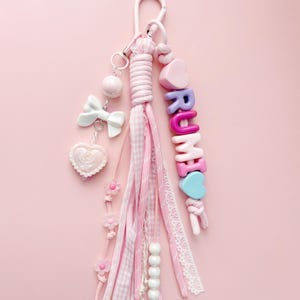May include: A pink keychain with a white clasp, adorned with pink and white ribbons, beads, and charms. The charms include a bow, a heart, and the name "RUMI" spelled out with colourful letter beads. The background is a solid pink.